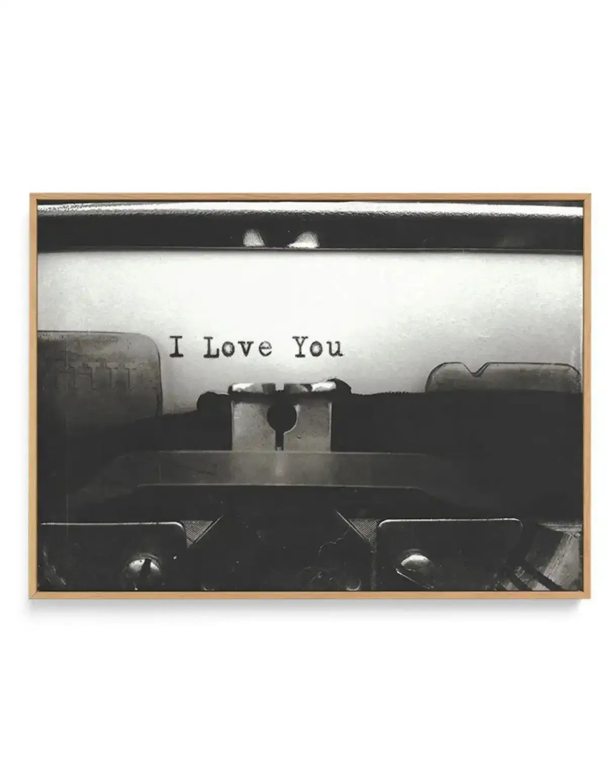 Black and white photograph of a typewriter shot from above, showing the paper roller and keys in sharp detail. The typed text "I Love You" appears on white paper in the center. The image has high contrast with deep blacks and bright whites, creating a vintage aesthetic. The photograph is mounted on a canvas print and housed in a natural wood frame with a white border.