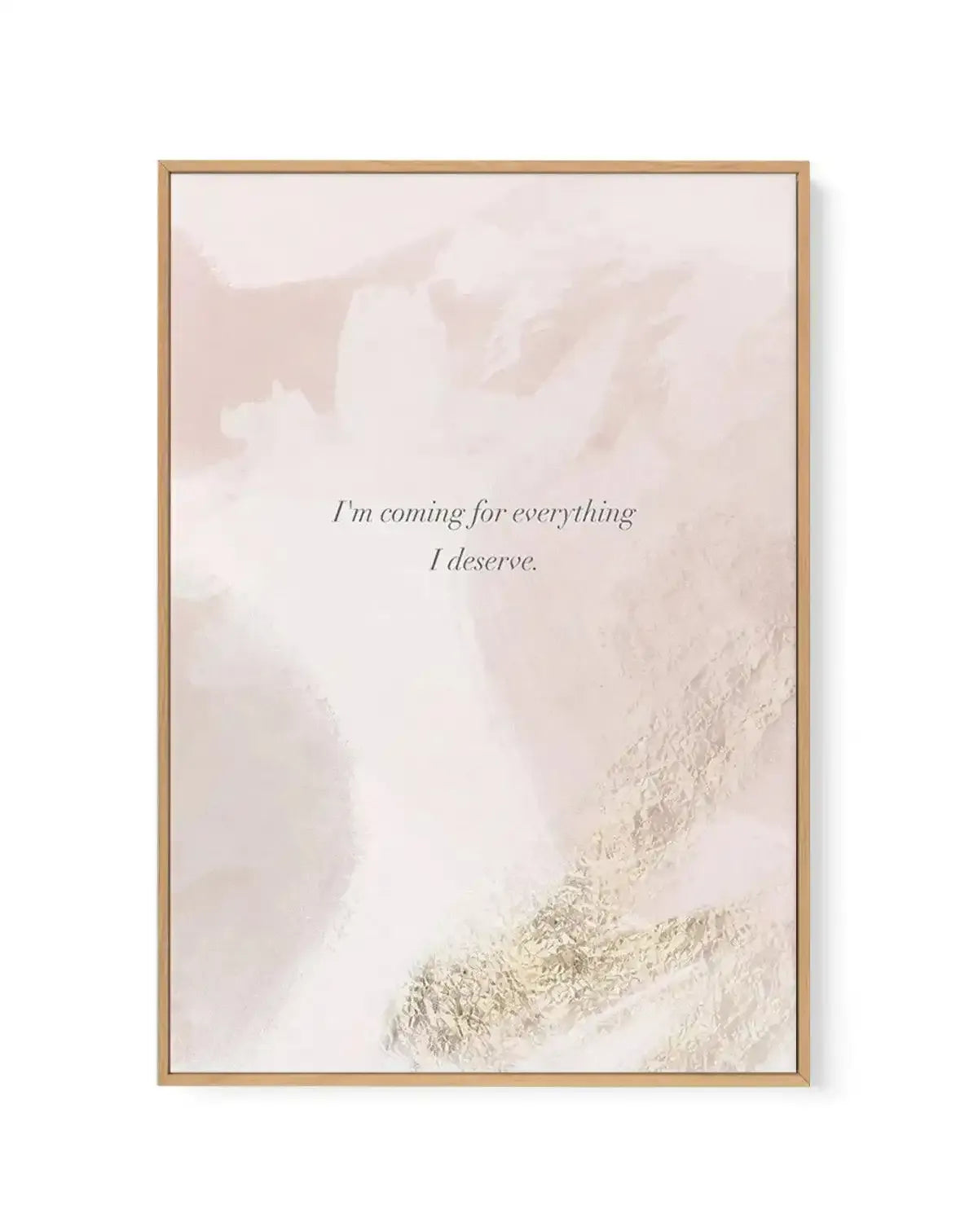 Framed canvas art print with a natural wood frame featuring an abstract landscape photograph in muted beige and cream tones. Snow-covered mountainside or terrain visible in the lower right corner with soft, blurred peaks in the background. Centered in italicized serif font is the affirmation text "I'm coming for everything I deserve" in dark gray. The overall composition has a serene, minimalist aesthetic with a soft, dreamy quality enhanced by the neutral color palette and photographic texture.
