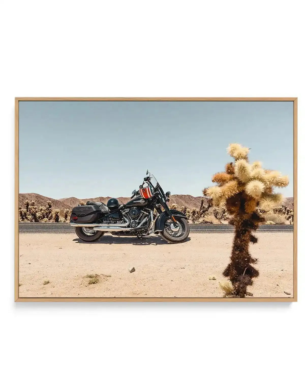 Framed canvas art print featuring a desert landscape photograph with a black cruiser motorcycle parked on sandy ground in the center. A distinctive Joshua tree with pale and golden-brown spiky branches frames the right foreground. Arid mountains and scattered desert vegetation create the background beneath a clear blue sky. The photograph has warm, natural tones with beige sand, deep blacks in the motorcycle, and muted earth colors. Presented in a natural wood frame with clean lines.