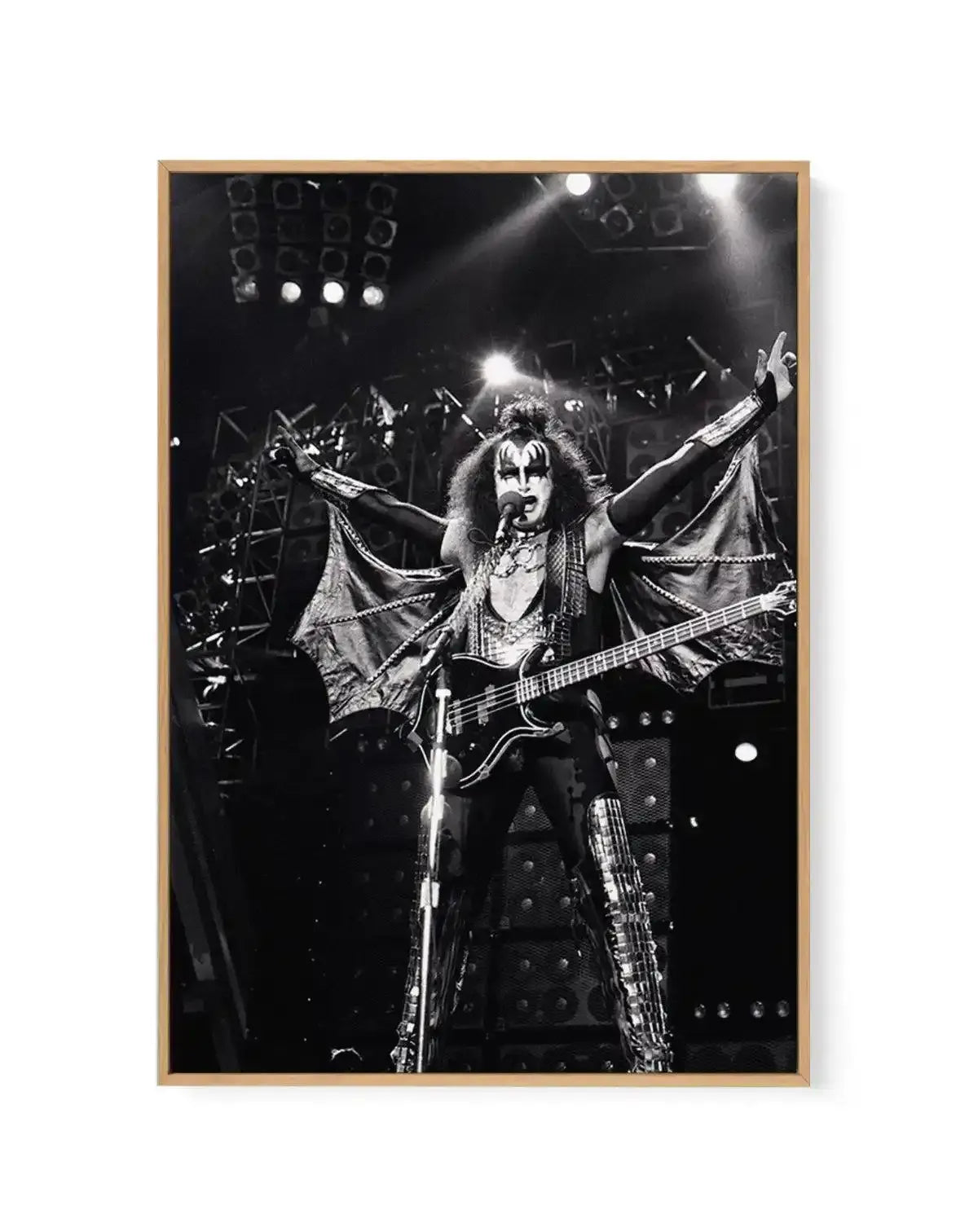 Black and white framed canvas art print featuring a dramatic live performance photograph of a rock musician with face paint and long hair, wearing an ornate studded costume with wing-like fabric elements, playing bass guitar on stage with arms raised. Stage lighting and amplifiers visible in the background. Image mounted in natural wood frame with white border.