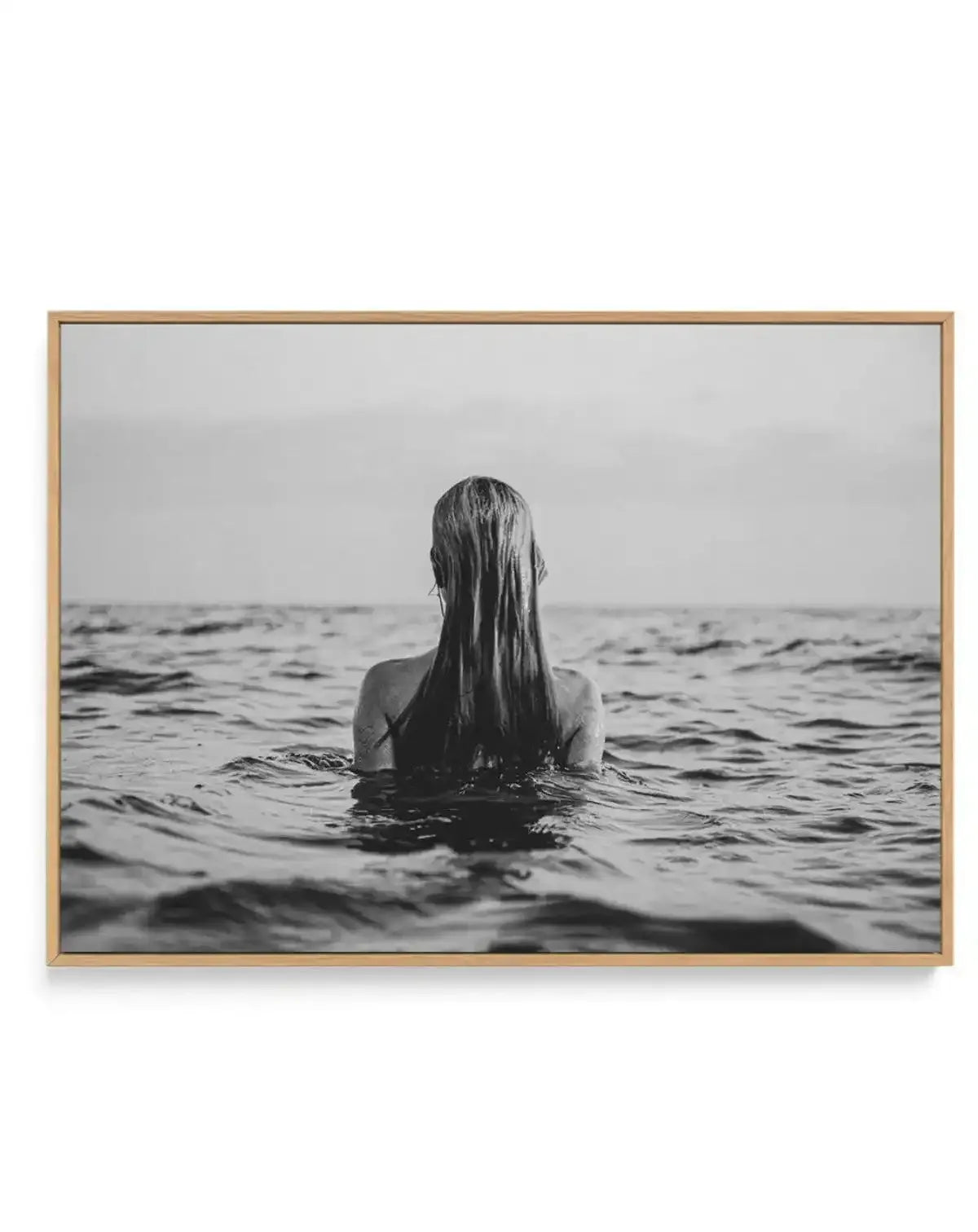 Framed black and white photograph of a woman with long wet hair, seen from behind, standing in the ocean with gentle waves around her, displayed in a natural oak frame with white border.