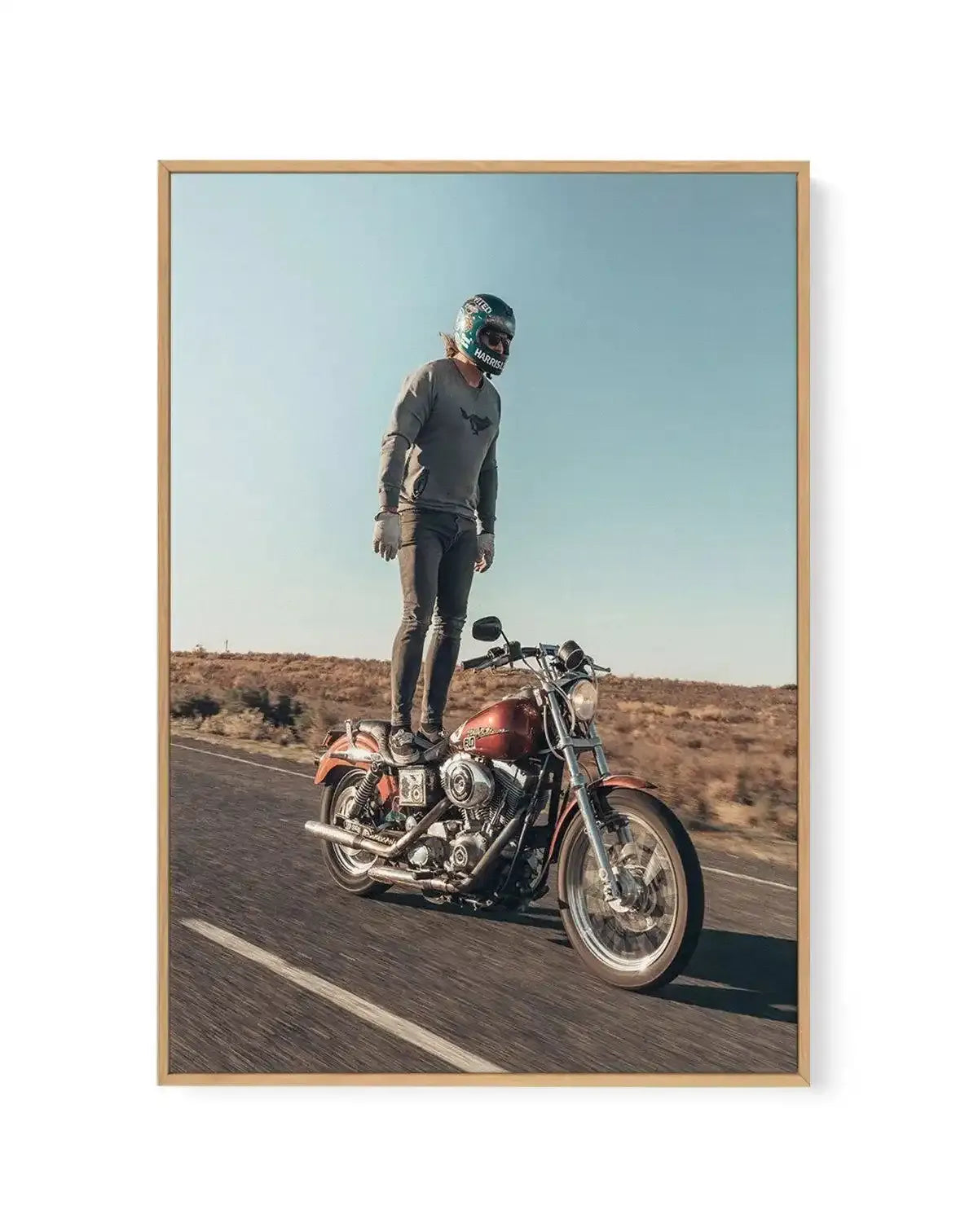 Framed canvas art print featuring a man standing on a moving red motorcycle on a desert road, with a clear blue sky. The man wears a grey long-sleeved shirt, dark pants, white gloves, and a dark helmet with "HARRIS" visible. The motorcycle is a classic style with chrome accents. The road has a white line, and the background shows dry, sparse vegetation under a bright sky. The print is displayed in a natural wood frame.