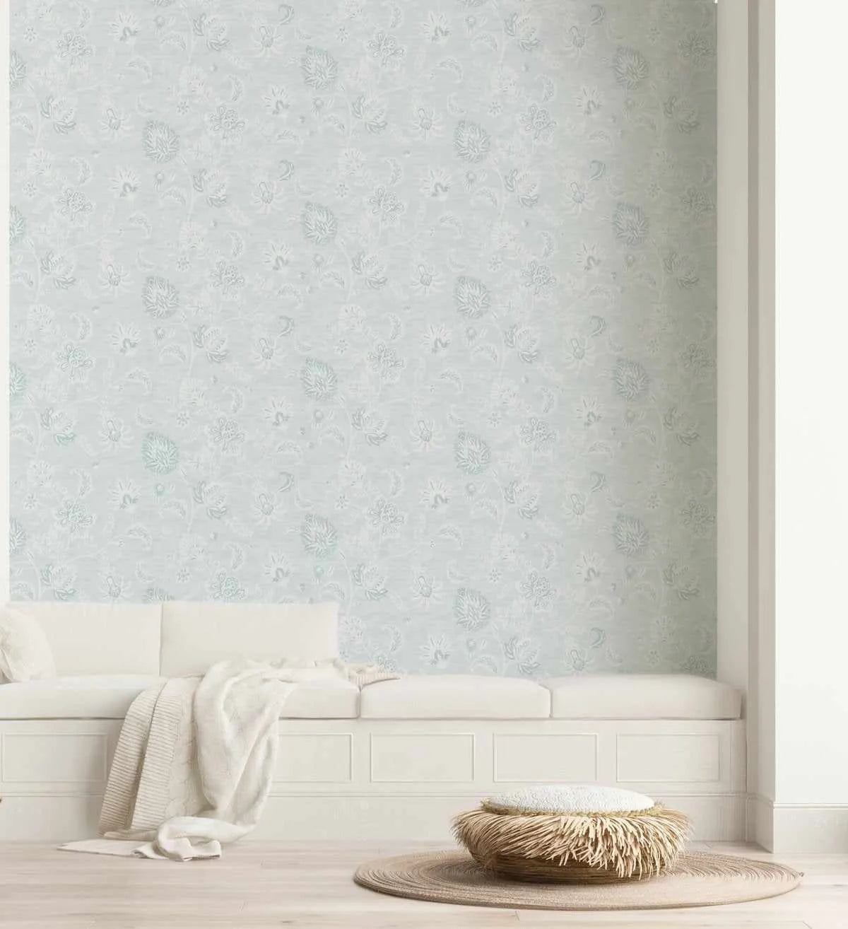 Lifestyle shot of a living room featuring Marsielle in Duck Egg Blue Wallpaper. The wallpaper has a repeating pattern of delicate, hand-drawn floral and botanical motifs in a soft duck egg blue and white on a pale grey-blue background. The room includes a white built-in bench with white cushions and a cream throw, and a round woven rug with a textured pouf on a light wooden floor.