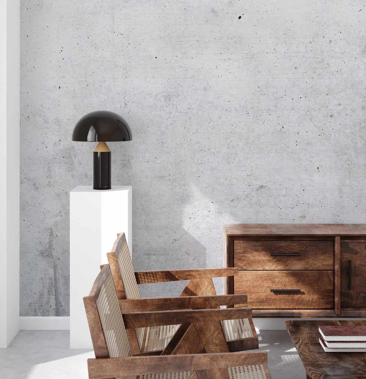 Lifestyle shot of a modern industrial living room featuring a concrete wallpaper with subtle grey tones and visible aggregate texture. The wallpaper serves as a backdrop for a black mushroom-shaped table lamp on a white pedestal, and two wooden armchairs with woven cane backs. A wooden sideboard and coffee table with books are also visible, bathed in natural light creating shadows on the wall and floor.