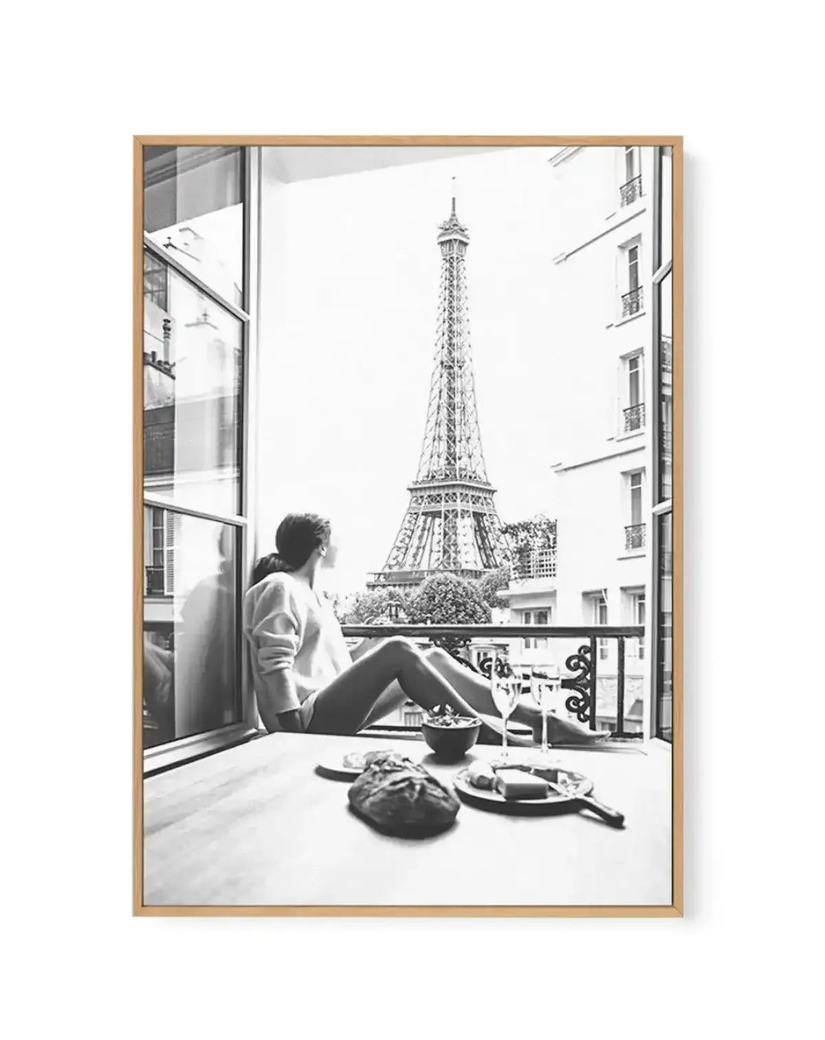 Black and white framed canvas art print titled "Mondays In Paris II" featuring a woman sitting on a window seat looking out at the Eiffel Tower. The photograph shows an open window with metal railings on a balcony, Parisian apartment buildings visible on either side, and a plate with food and bowl on the windowsill in the foreground. The image has a classic, nostalgic quality with soft grey tones throughout. The canvas is mounted in a natural wood frame with a white border.