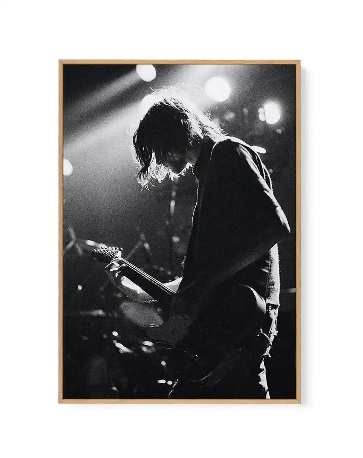 Framed black and white canvas print of a musician playing guitar on stage under bright spotlights. The musician, with long, dark hair, is silhouetted against the light, facing right and looking down at their guitar. The image captures a dynamic moment with visible light rays and a grainy texture, displayed in a natural oak frame.