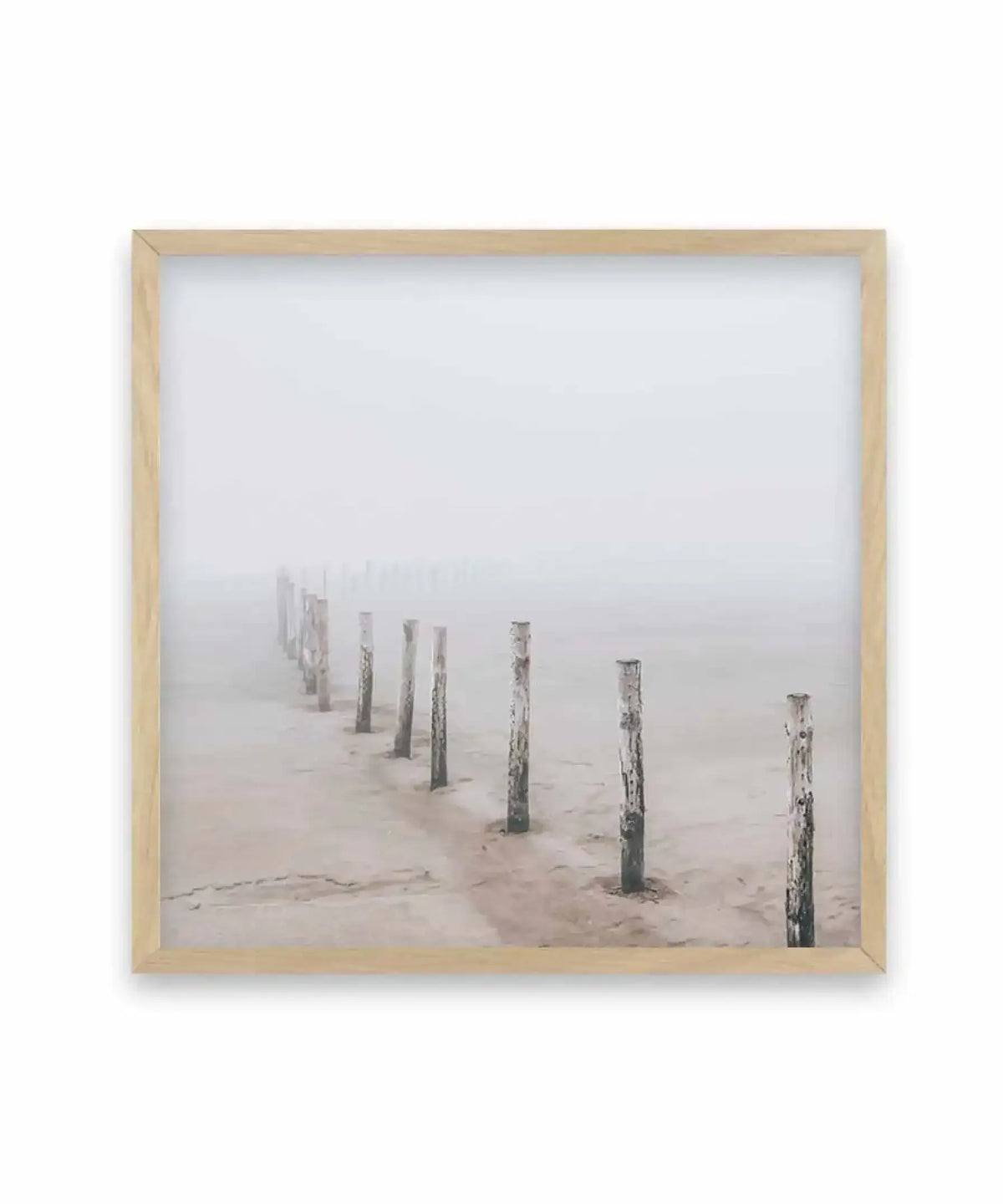 Nordic Seaside Square Art Print in natural wood frame showing a misty beach landscape. Weathered wooden posts recede into dense fog along sandy shore in muted tones of gray, beige, and soft taupe. Minimalist composition with moody, serene atmosphere. Framed in light natural wood with white border.