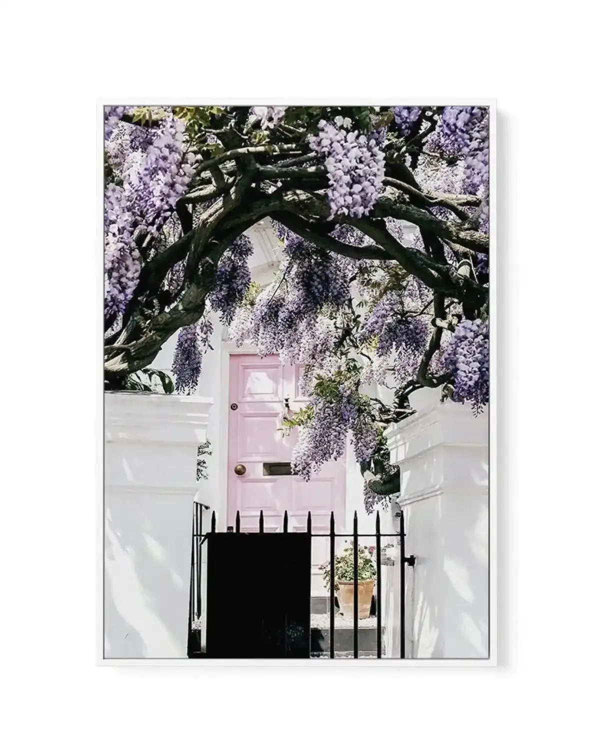 Framed canvas art print featuring a photograph of a white townhouse with a pale pink front door beneath an ornate black iron gate, all framed by cascading wisteria with lavender-purple flowers and dark twisted branches. The composition captures the romantic charm of a Notting Hill London street facade. The canvas is mounted in a white frame with white border.