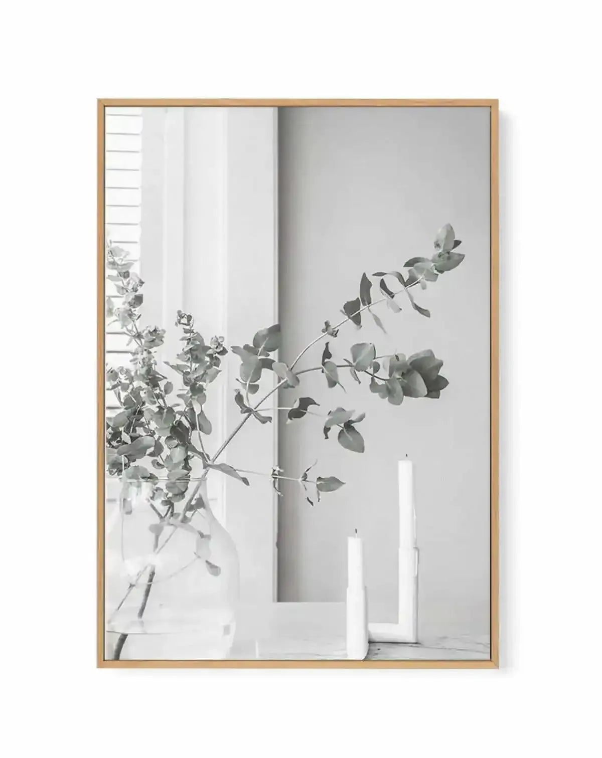 Framed black and white photograph of eucalyptus branches in a glass vase with two white candles, displayed in a natural oak frame with white border. The composition features a clear glass vase holding several eucalyptus branches with round leaves, positioned on a light-coloured surface next to two tall white pillar candles. The background shows a window with blinds on the left and a plain wall on the right, creating a minimalist and serene mood.