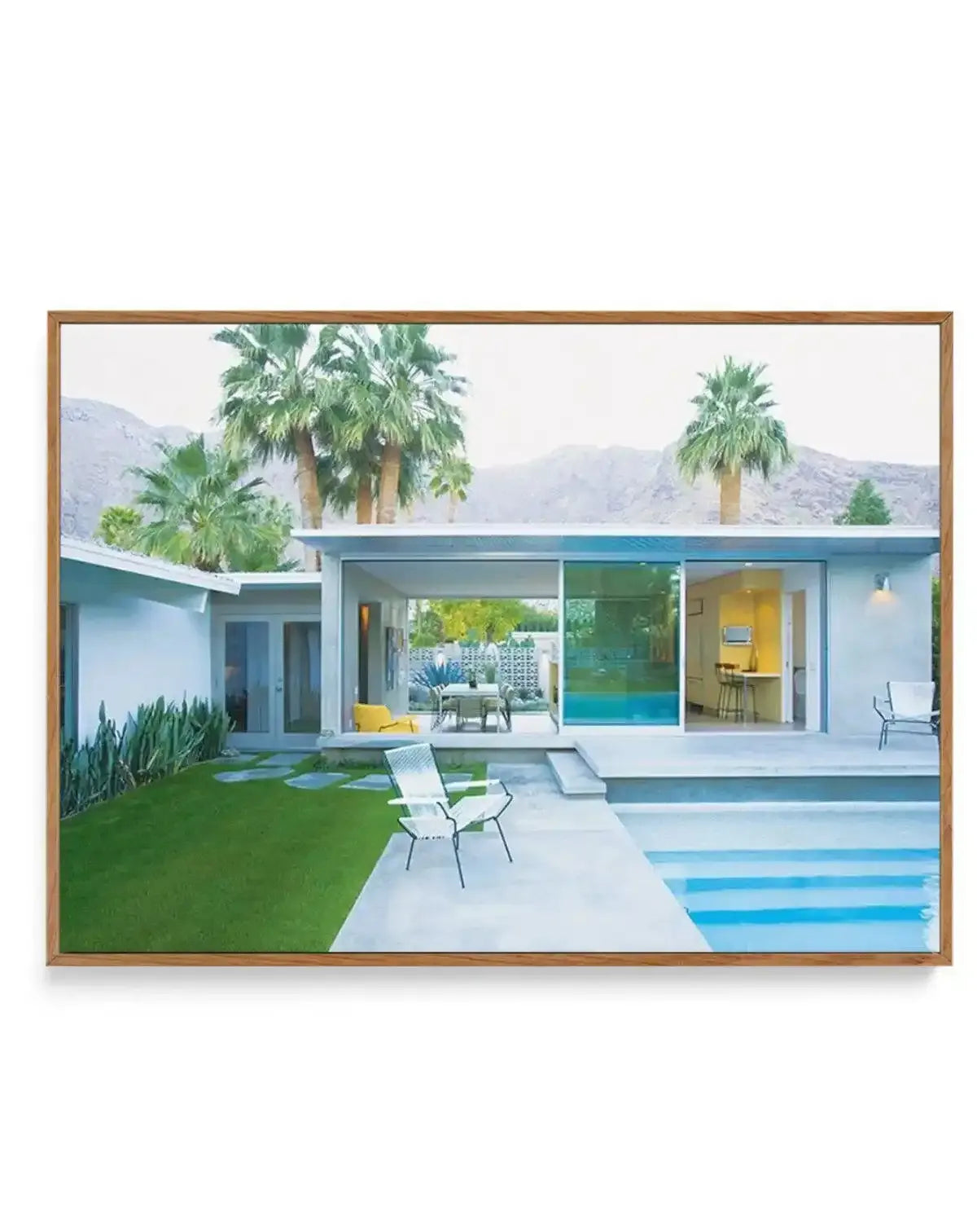 Framed canvas print of a mid-century modern house with a swimming pool and palm trees under a light sky, with a natural wood frame. The house is white with large glass sliding doors, revealing an outdoor dining area and an indoor kitchen. A white lounge chair sits by the pool, and mountains are visible in the background.