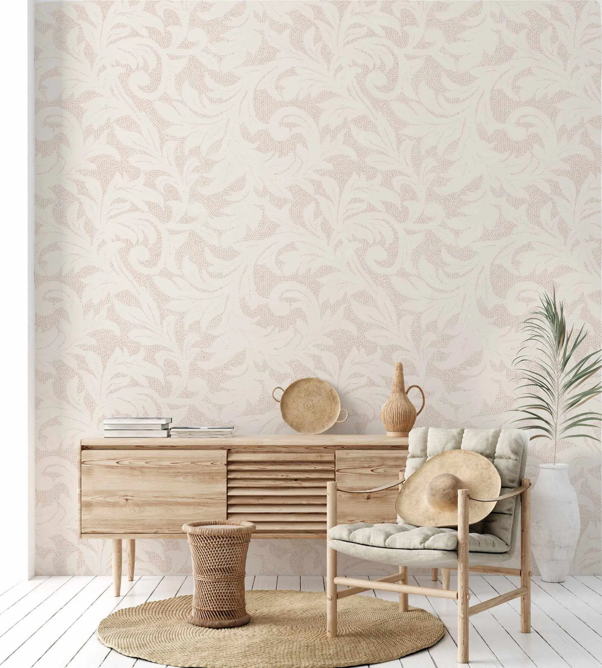 Lifestyle shot of a living room featuring Provence in Sand wallpaper. The wallpaper has a repeating pattern of organic, swirling leaves and vines in a subtle, light beige tone against an off-white background, with a textured, dotted effect within the leaf shapes. The room includes a light wood sideboard with books, a woven basket, a round jute rug, a light wood chair with a straw hat, a large white vase with a green plant, and a decorative ceramic jug.