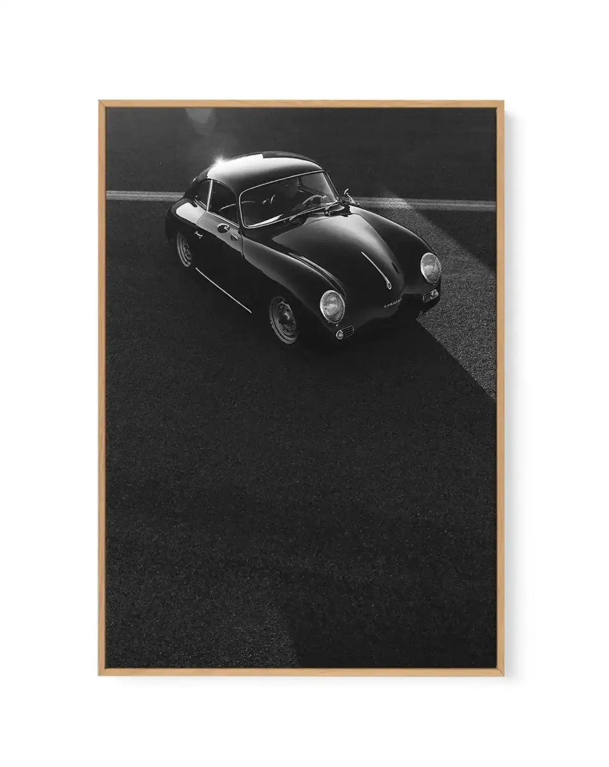 Black and white overhead photograph of a vintage Porsche 356 sports car positioned on dark asphalt with a white lane line visible, mounted on a framed canvas with a natural wood border and white mat.