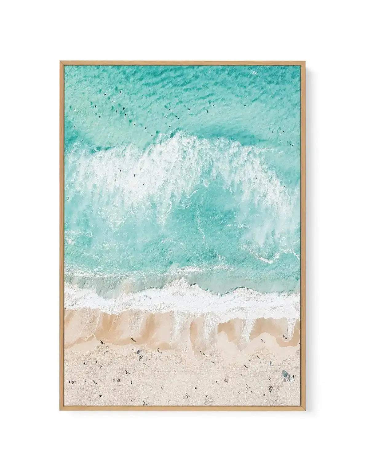 "Gallery-wrapped canvas print of an aerial photograph of a beach scene, framed in natural wood. The composition features vibrant turquoise ocean water with white foamy waves breaking onto a wide expanse of light beige sand. Numerous small dark figures, likely people, are scattered across the sand and in the shallow water, suggesting a lively beach atmosphere."