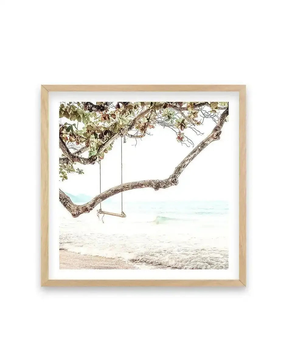 Framed photograph of a serene beach scene with a wooden swing hanging from a tree branch, displayed in a natural oak frame with white border. The composition features a gnarled tree branch with green and reddish-brown leaves extending over a light sandy beach with gentle white waves breaking on the shore, and a pale blue ocean under a bright sky.
