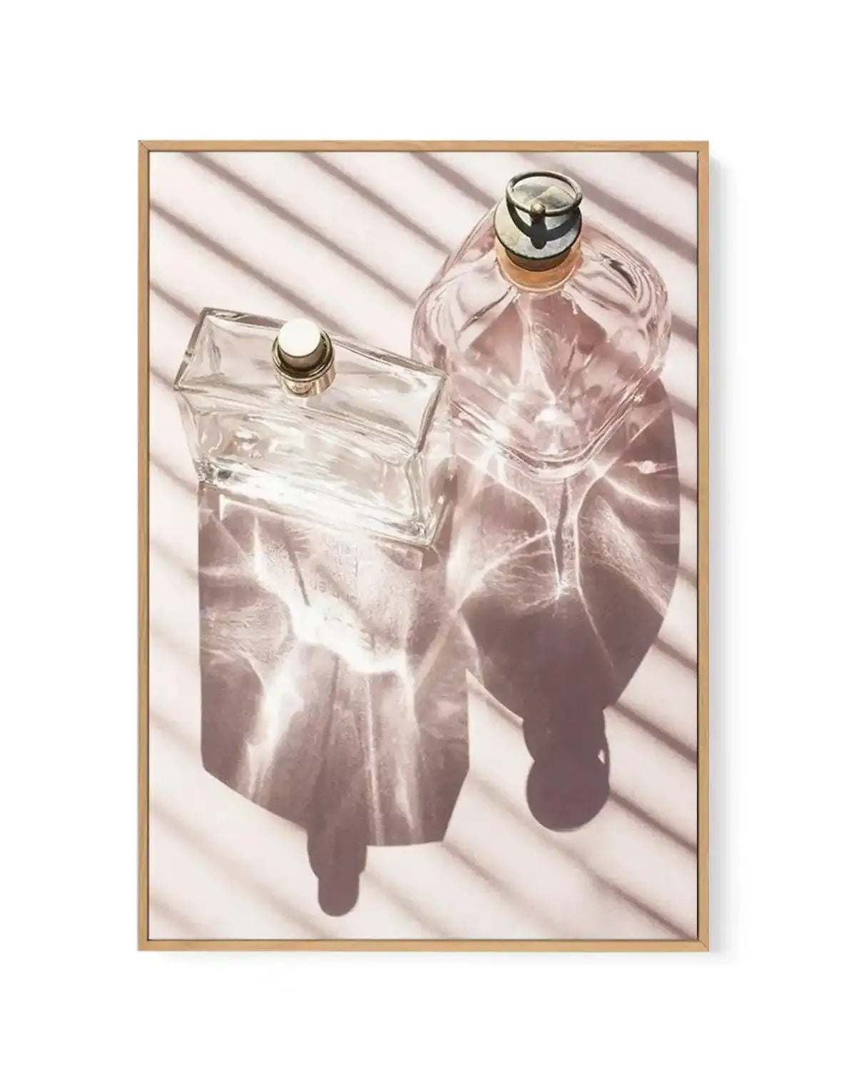 Framed canvas art print featuring two glass perfume bottles photographed from above against a white background with diagonal shadow patterns. The left bottle is a clear rectangular flacon with a gold-trimmed stopper, while the right bottle is a rounded shape filled with pale rose-tinted liquid, also topped with a gold and black cap. Soft shadows cast across the composition create depth and dimension. The artwork is mounted in a natural wood frame with a white border.