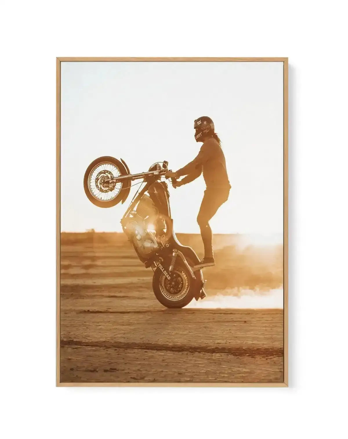 "Framed canvas print of a person performing a wheelie on a motorcycle in a desert landscape at sunset, displayed in a natural wood frame with a white border. The photograph captures the rider standing on the motorcycle, with the front wheel lifted high, and dust trailing from the rear wheel. The warm, golden light of the setting sun creates a dramatic silhouette effect, highlighting the rider's helmet and the motorcycle's details against a bright, hazy sky and a flat, sandy ground."