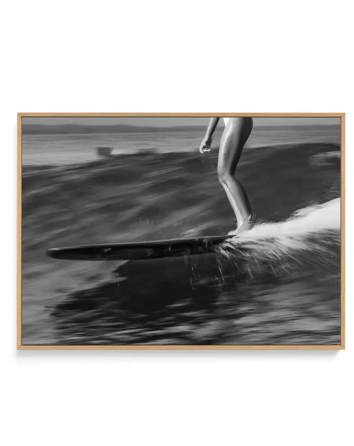 Framed black and white photograph of a surfer riding a wave in Malibu, displayed in a natural oak frame with white border. The composition captures the surfer's lower body and board as they glide across the water, creating a spray of white foam. The ocean stretches to the horizon under an overcast sky, with a sense of motion conveyed by the blurred water.