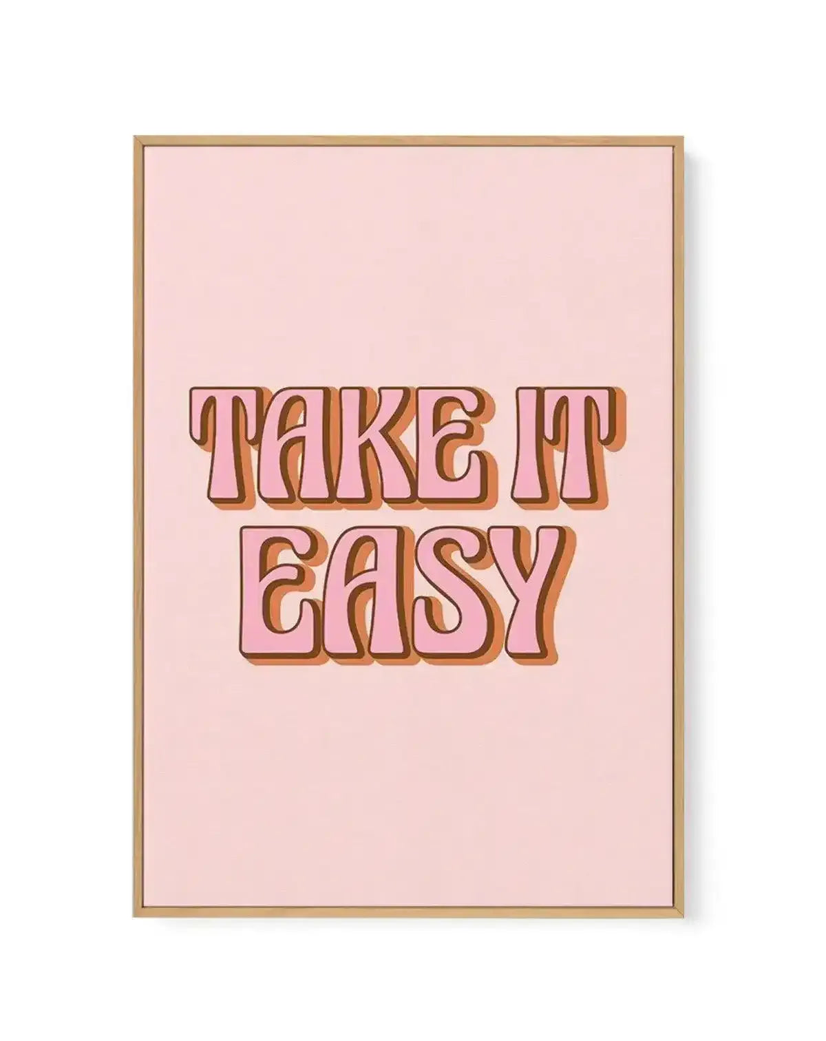 Framed canvas art print featuring the text "TAKE IT EASY" in large, bold, retro-style lettering with orange and brown gradient shading and dark burgundy outlines. The text is centered on a soft blush pink background. The canvas is surrounded by a natural wood-tone frame with a white border, creating a clean, minimalist presentation against a white background.
