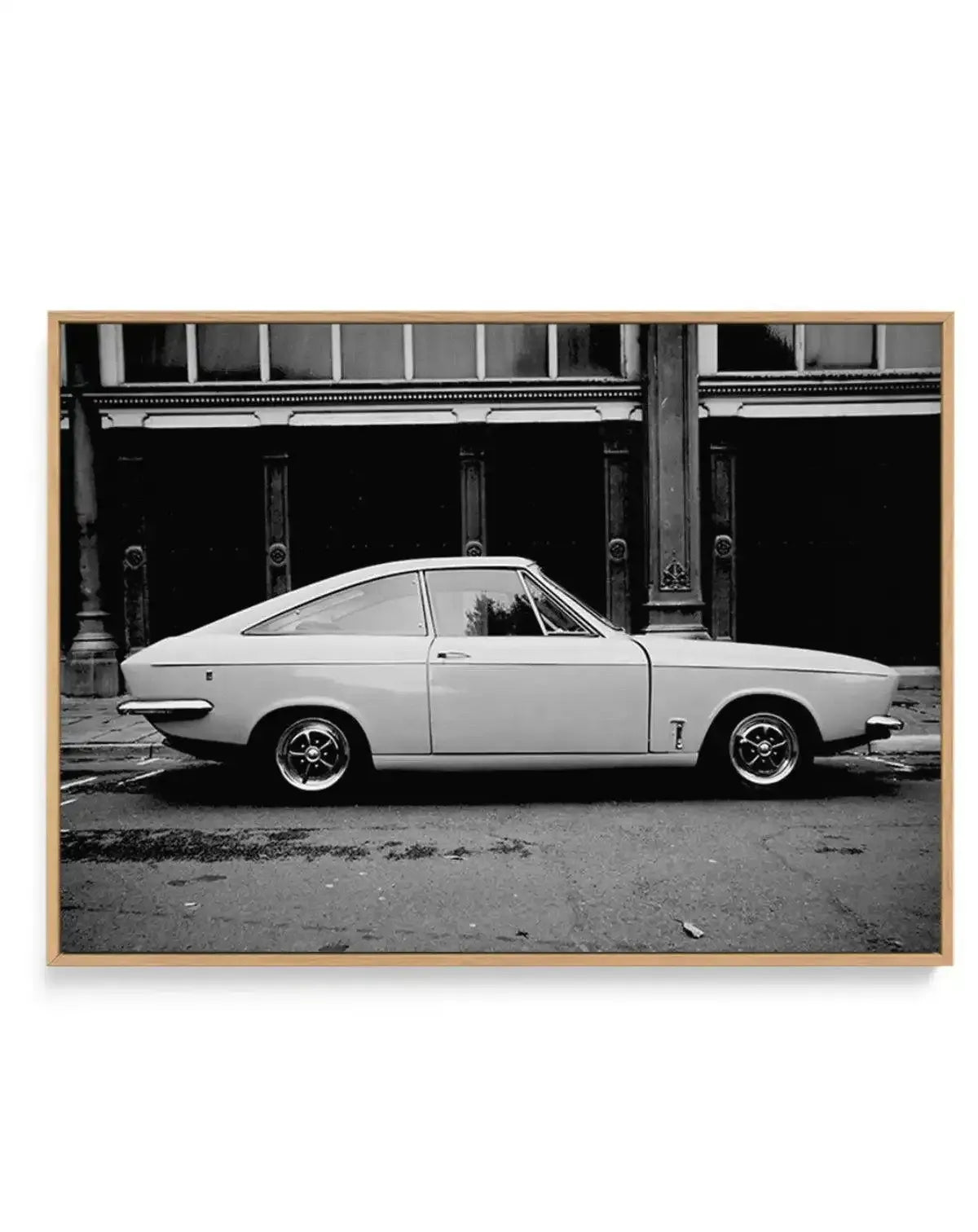 Black and white photograph of a vintage Bond Equipe sports car parked on a street, shown in profile view with the driver's door open. The car features a sleek, curved coupe body with chrome detailing and wire-spoke wheels. Behind the vehicle is a storefront with dark windows and ornate architectural columns. The photograph is printed on canvas and framed in light natural wood, displayed against a white background.