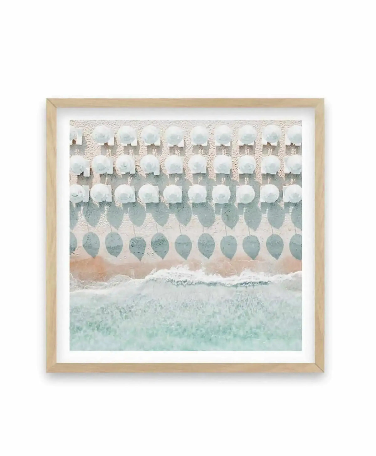 White Umbrellas Square Art Print in natural wood frame with white border, featuring a soft watercolor-style illustration of rows of white umbrellas arranged in a grid pattern against a beach scene with turquoise shallow water, sandy beach, and cream-colored textured wall background in muted tones of white, gray, sage green, and soft peach.