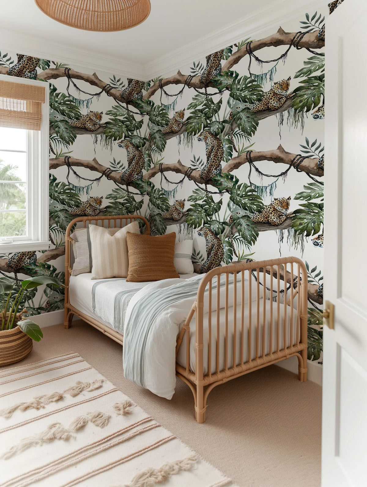 Lifestyle shot of a child's bedroom featuring a wallpaper with a repeating pattern of leopards lounging on tree branches amidst lush green jungle foliage. The wallpaper covers all visible walls, creating an immersive tropical theme. The room includes a light-colored rattan daybed with white and striped pillows, a light blue and white striped duvet, and a woven rug with cream stripes and tassels on a light beige carpet. A potted plant in a woven basket sits near a window with a bamboo blind.