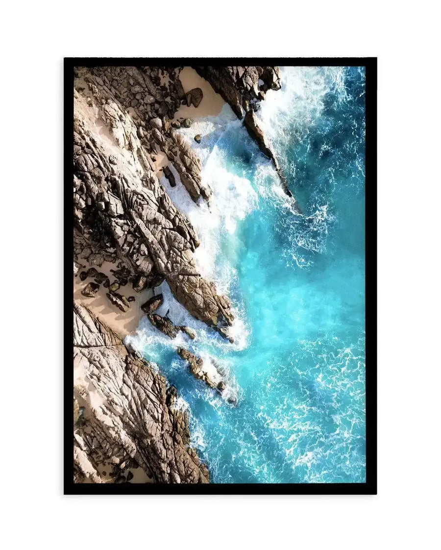 Framed aerial photograph of a coastal beach scene with a black frame and white border. The composition features rugged, light brown rock formations and small sandy coves meeting vibrant turquoise ocean water with white foamy waves crashing against the shore.