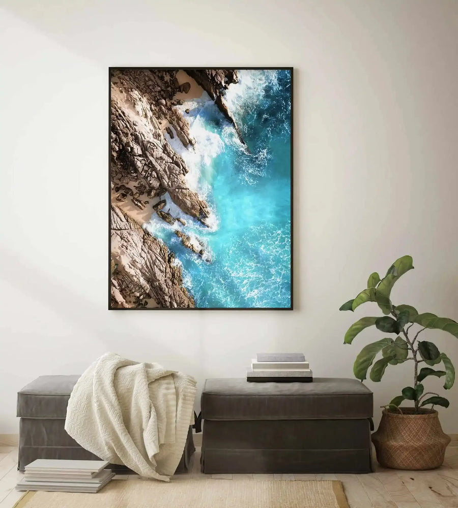 Lifestyle shot of a minimalist living room featuring a large framed art print of an aerial beach scene with natural black frame and white border. The print depicts turquoise ocean water crashing against a sandy beach with large, textured rocks. Below the print are two dark grey upholstered ottomans, one with a cream textured throw blanket and a stack of books, and the other with a potted plant in a woven basket.