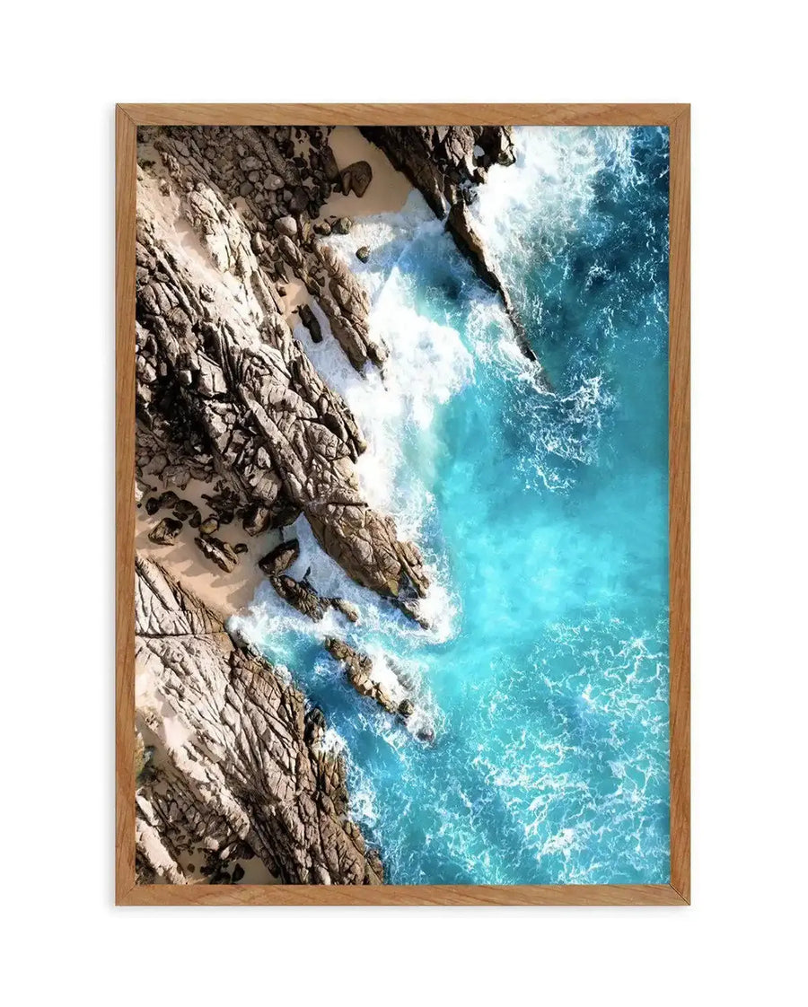 Framed aerial photograph of a coastal scene with natural wood frame and white border. The composition features a rugged coastline with sandy coves and turquoise ocean water crashing against the rocks, creating white foam.