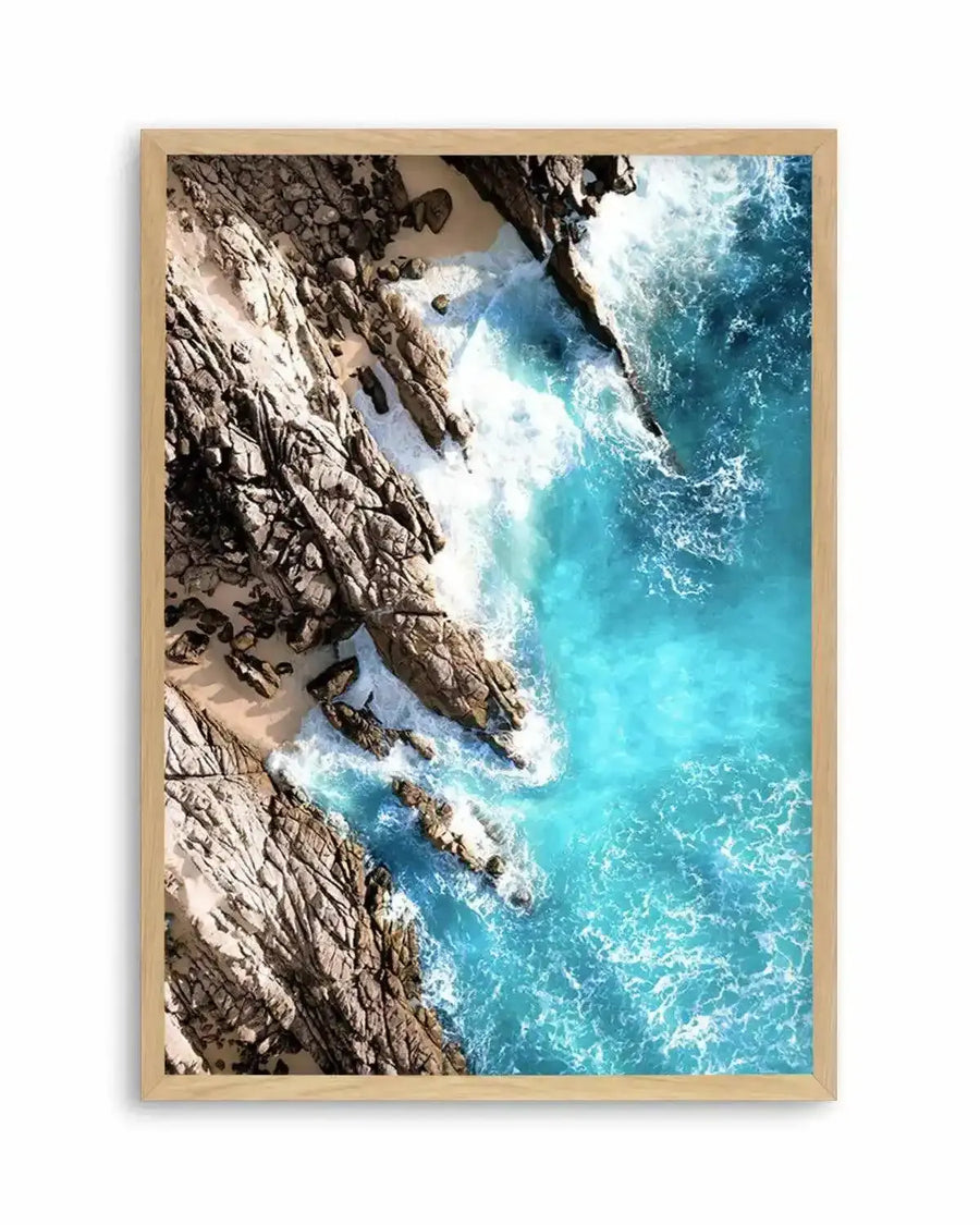 Framed aerial photograph of a coastal scene with a natural wood frame and white border. The composition features rugged rock formations and small sandy coves meeting vibrant turquoise ocean water with white foamy waves crashing against the shore.