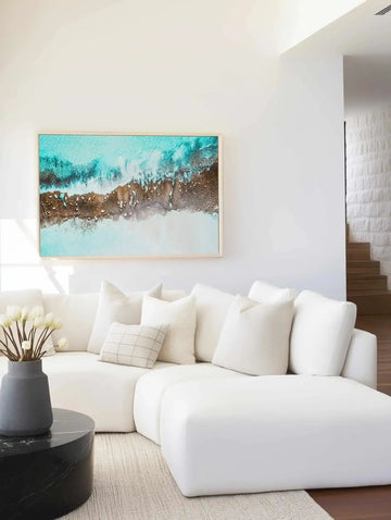Turquoise and brown aerial coastal print in natural wood frame, above a white sofa in a bright living room