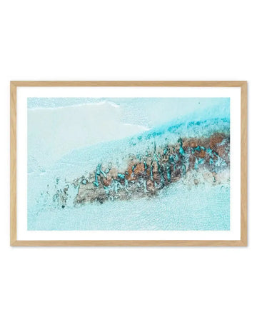 Framed aerial photograph of 11 Mile Beach, Esperance, with a natural wood frame and white border. The image captures the turquoise shallow waters of the ocean meeting a white sandy beach, with a prominent band of dark brown and teal-coloured coral reef running diagonally across the frame. The water is clear, revealing the sandy bottom and the intricate textures of the reef.