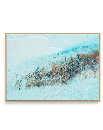 Aerial photograph canvas print of a beach scene with clear turquoise water and a strip of white sand, framed in natural oak. The composition captures a shallow reef or rock formation in the water, appearing as a dark brown and teal mass with organic shapes, creating a striking contrast against the light blue water.