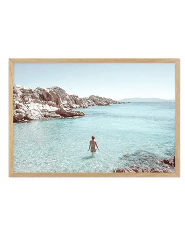 Framed photograph of a serene coastal scene with a lone person wading into clear turquoise water, displayed in a natural oak frame with white border. The composition features rugged, light-coloured rock formations lining the shore under a pale blue sky, with distant mountains on the horizon.