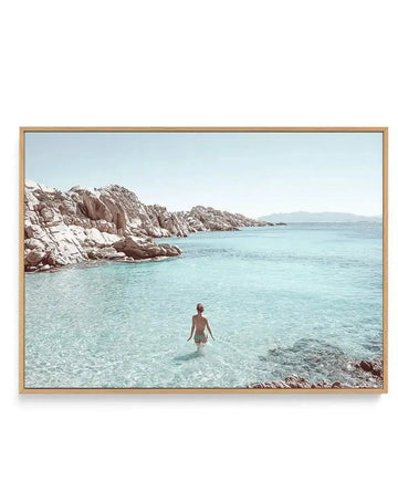 Woman in bikini wading in clear turquoise rocky cove, pale blue sky, on canvas in a natural wood float frame