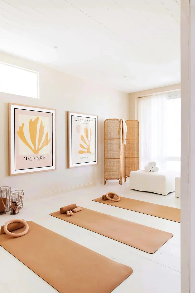 Abstract art print titled "Papiers Decoupes III" displayed on a white wall in a natural wood frame with white border. The artwork features minimalist golden-yellow organic shapes and curved forms on a cream background with soft pink accents. Hung in a bright, minimalist yoga studio with blonde wood furnishings, tan yoga mats, wooden props, and a woven room divider.