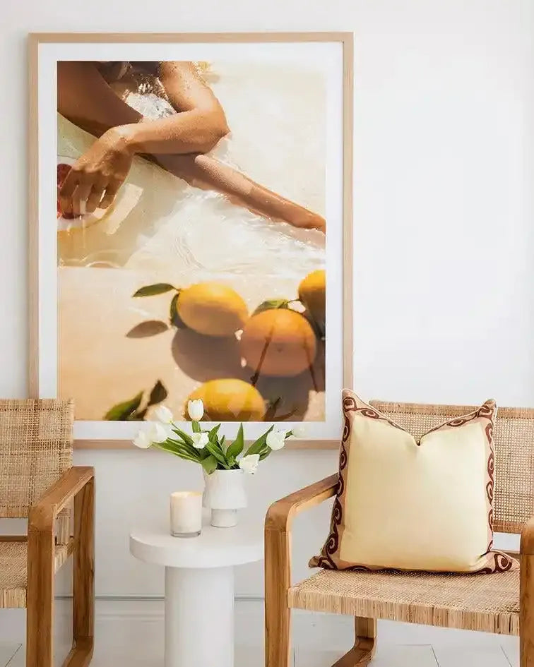 Lifestyle shot of a framed art print displayed in a bright room with two cane chairs and a small white side table. The print is a photograph of a person's arms and legs partially submerged in clear water, with several oranges and lemons floating on the surface. The water is light and reflective, and the fruit casts soft shadows. The frame is natural wood with a white border. On the side table are a white vase with white tulips and a lit candle.