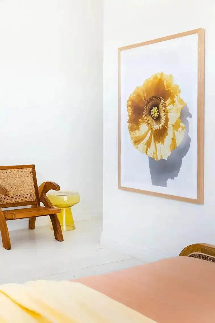 Yellow Poppy Art Print displayed in a light wood frame with white border, hung on a white wall in a bright room. The artwork features a large yellow poppy flower with delicate, layered petals and a dark brown center, depicted in soft watercolor style against a light background with a subtle gray shadow effect. Below the framed print sits a wooden chair with woven rattan backing and a yellow cylindrical side table.