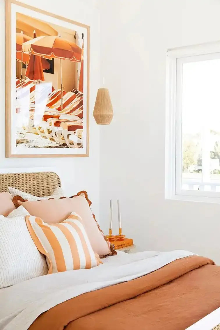 Framed art print displayed above a bedroom headboard showing a vintage beach scene with orange and cream striped umbrellas, loungers, and beach chairs on sand. The photograph has warm golden and terracotta tones and is mounted in a light wood frame with white border. The bedroom features coordinating orange and white striped pillows and rust-colored bedding, complemented by a woven rattan headboard and wooden pendant light.