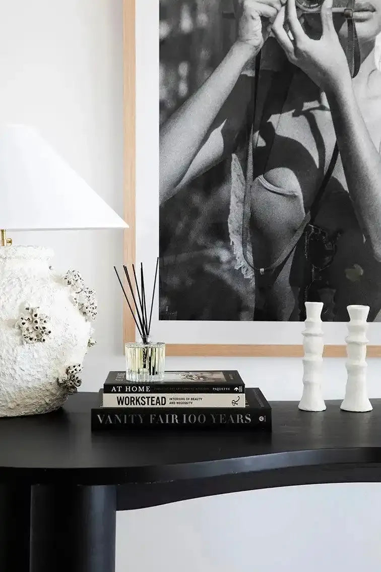 Black and white photographic art print displayed on a white wall in a natural wood frame, showing a close-up of a woman's torso and arms holding a vintage camera. The photograph has a grainy, documentary quality typical of mid-century photography. Below the framed print sits a black table styled with stacked design books including "Vanity Fair 100 Years" and "Workstead," a clear glass reed diffuser with black reeds, white ceramic sculptural vessels, and a textured white decorative object on the left side.