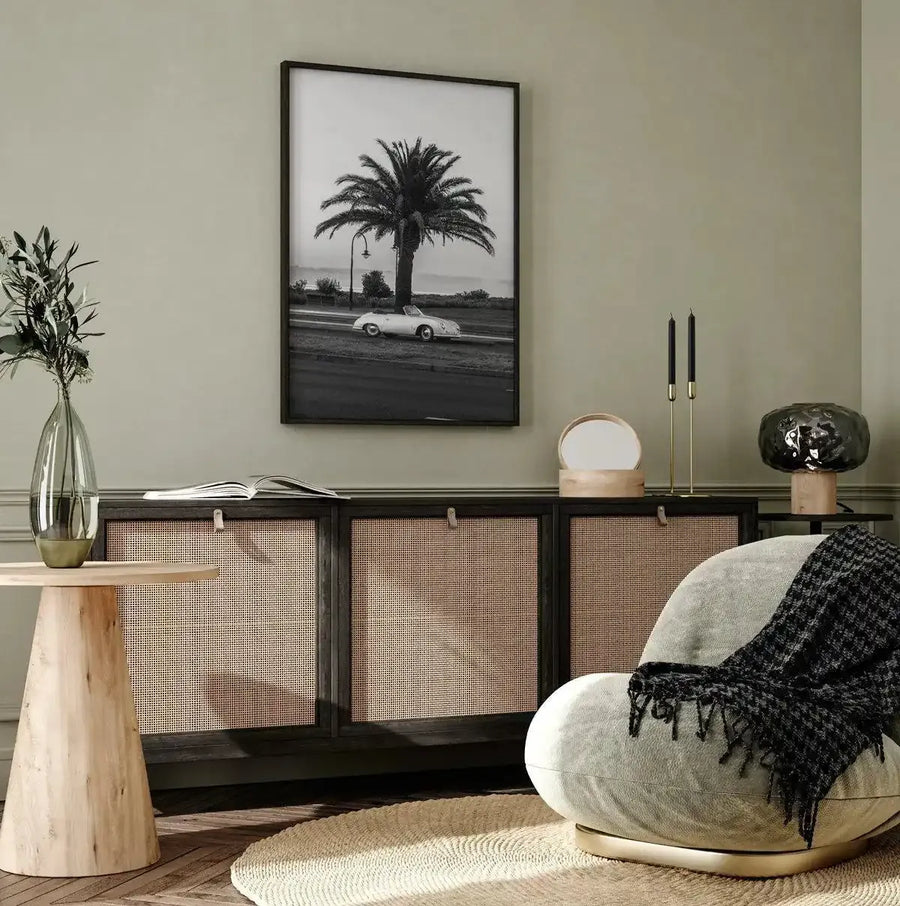 Lifestyle shot of a minimalist living room featuring a framed black and white photograph of a vintage white convertible car parked next to a palm tree on a roadside. The art print is displayed above a dark wood sideboard with cane paneling. The room includes a light grey armchair with a black and white patterned throw, a round wooden side table, and decorative items such as a vase with greenery, candles, and lamps.