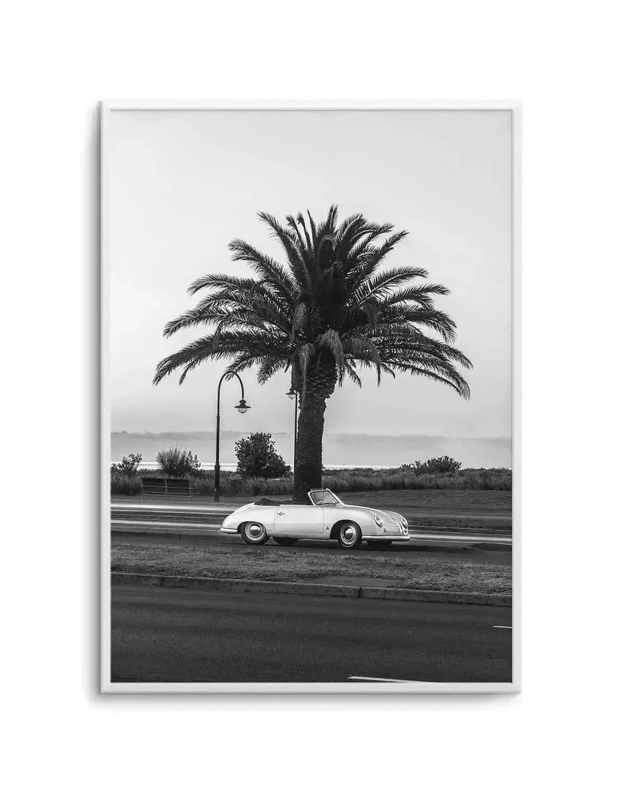 Framed black and white photograph of a vintage white convertible car parked on a grassy verge next to a road, beneath a tall palm tree. In the background, a body of water and a hazy horizon are visible under an overcast sky. The print is displayed in a white frame with a white border.