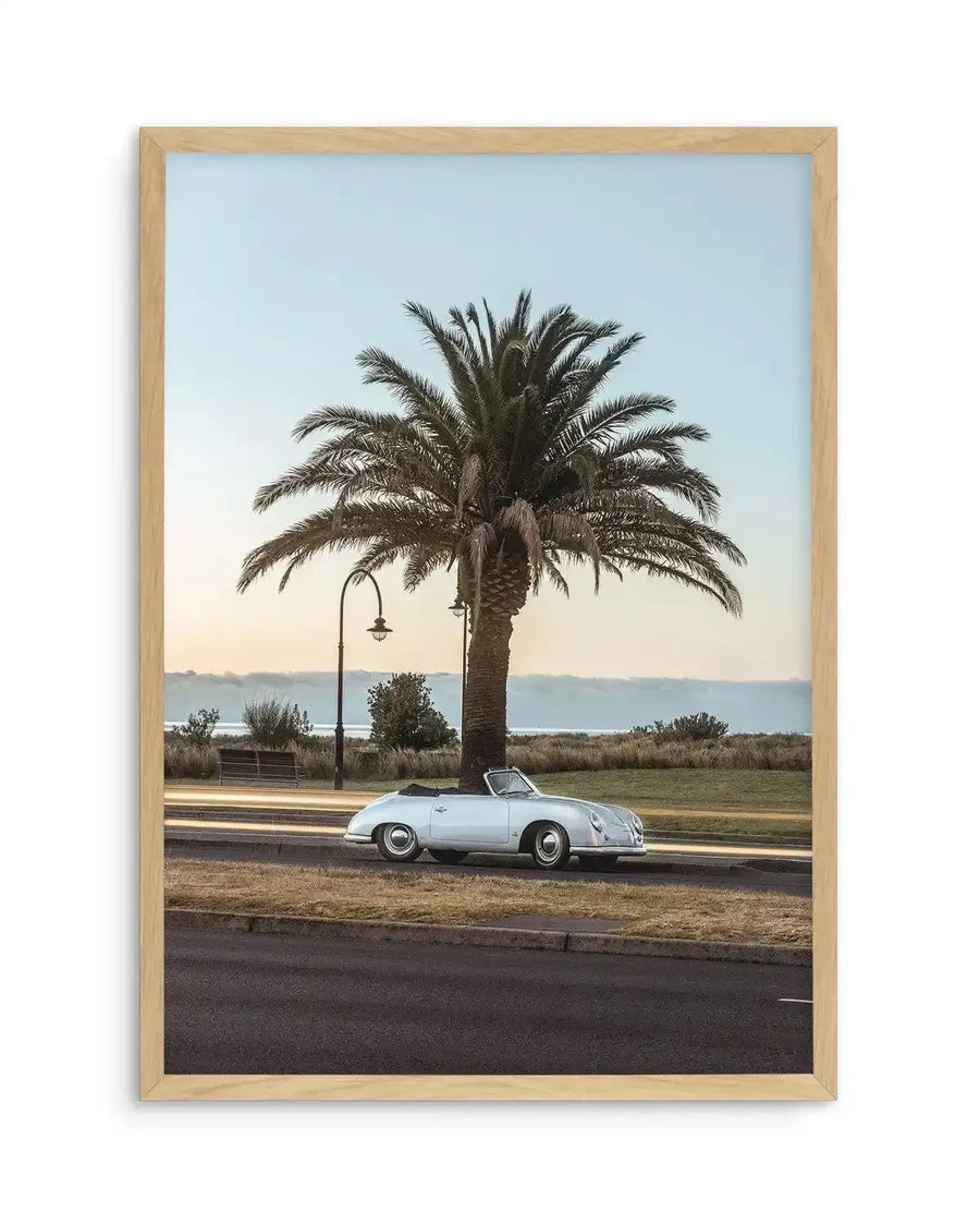 Framed photography art print featuring a vintage white convertible car parked on the side of a road next to a large palm tree, with the ocean visible in the background under a pale blue and orange sunset sky. The print is displayed in a natural oak frame with a white border.