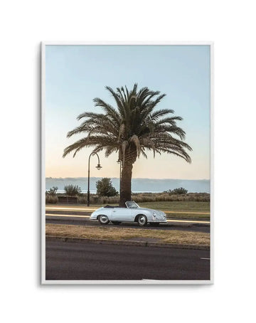 Framed photography art print capturing a classic white convertible car parked beside a palm tree at sunset, with a natural wood frame and white border. The composition features a vintage Porsche 356 parked on the roadside, with a tall palm tree and a street lamp behind it. In the background, a calm body of water meets a hazy horizon under a soft, light blue sky with warm golden light.