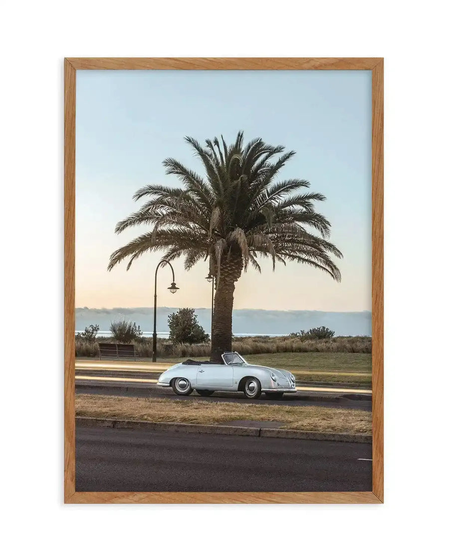 Framed photography art print featuring a vintage white convertible car parked on the side of a road next to a large palm tree at dusk, with a natural oak frame and white border. The composition captures the soft light of sunset over a distant body of water, with light trails visible on the road.