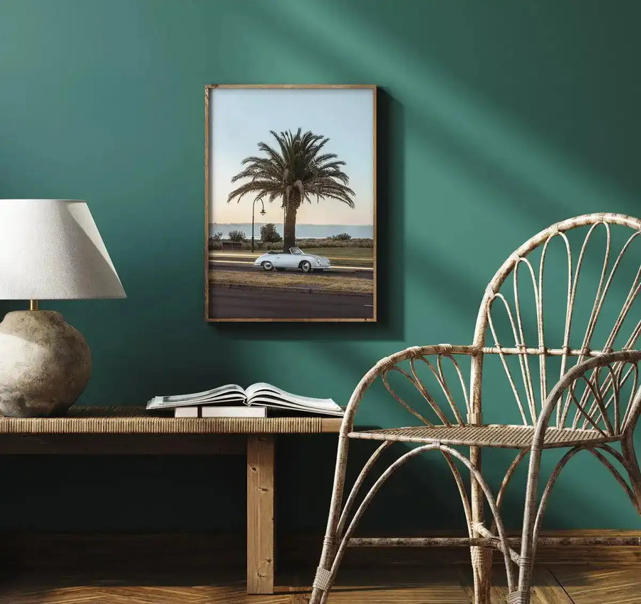 Lifestyle shot of an art print displayed on a dark teal wall in a room with natural wood furniture. The framed art print features a classic white convertible car parked on a road next to a large palm tree, with the ocean visible in the background under a soft, clear sky. The room includes a wooden bench with a book and a lamp, and a woven rattan chair, all bathed in warm sunlight.