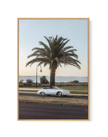 Framed canvas art print featuring a vintage white convertible sports car parked beneath a large palm tree on a coastal promenade. A street lamp stands nearby, with a wooden bench visible in the background. The scene overlooks a calm sea under a soft, hazy sky in muted blue and cream tones. The photograph has a nostalgic, mid-century aesthetic with natural lighting. The canvas is mounted in a natural wood frame with a white border.