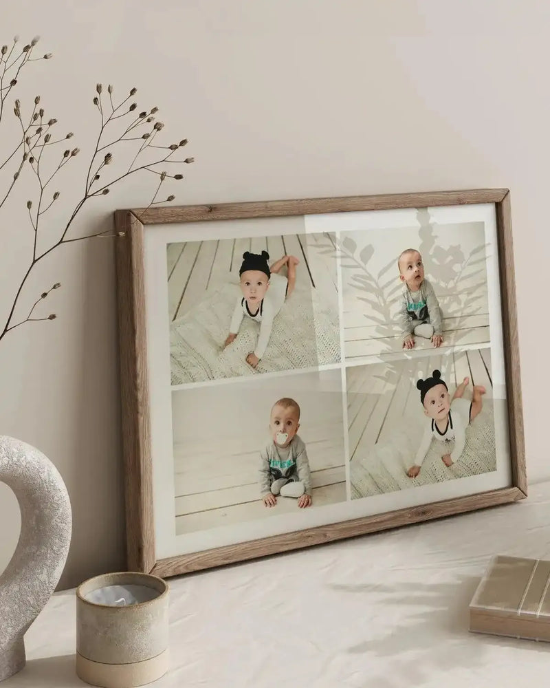 Lifestyle shot of a framed art print featuring four photographs of babies, displayed on a white table with a dried floral arrangement and a candle. The art print has a natural wood frame and white border, showcasing four individual images of babies in different poses and outfits, creating a collage effect.