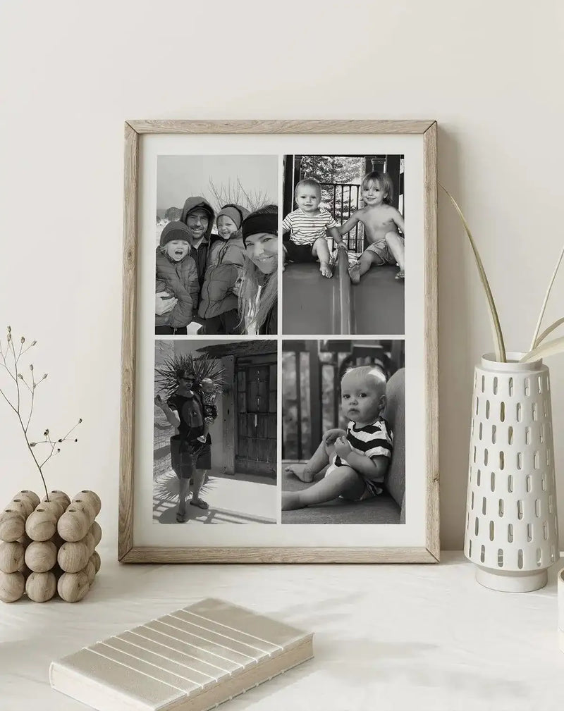 Framed black and white photography art print featuring four family portraits in a grid, displayed in a natural oak frame with white border. The top left photo shows a family of four in winter clothing, smiling. The top right shows two young children on a slide. The bottom left shows a man holding a baby outdoors. The bottom right shows a baby sitting on a couch. The print is on a white table next to a white vase with dried foliage and a decorative wooden object.