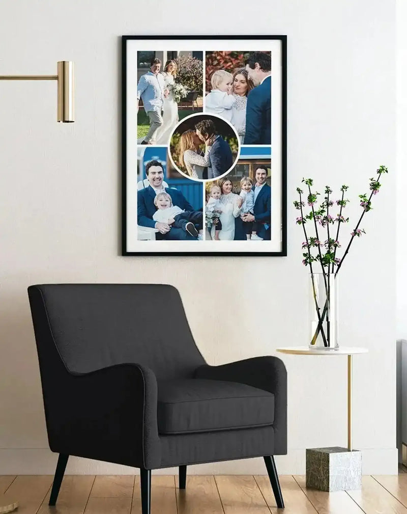 Large framed art print displayed above a dark grey armchair in a minimalist living room. The print is a collage of five family photographs, including a wedding couple, a couple with a baby, a father holding a child, and a family with two children. The central photo is a circular crop of a couple kissing. The print is displayed in a black frame with a white border. A gold-toned wall sconce is visible on the left, and a white side table with a vase of flowering branches is on the right.