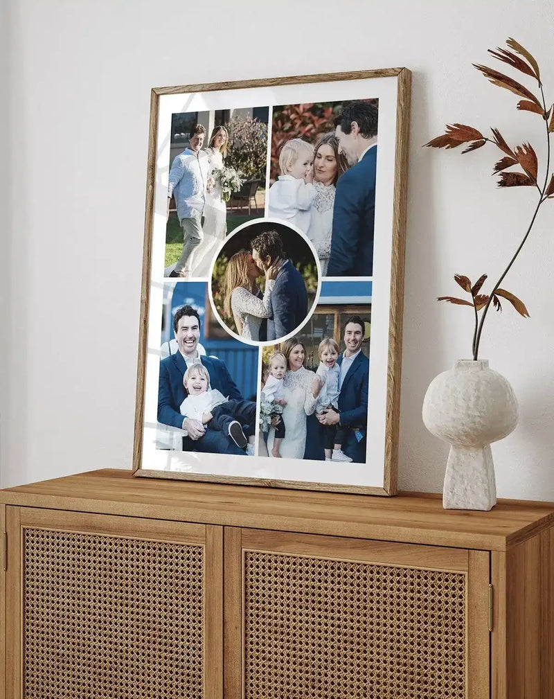 "Large framed collage art print displayed on a wooden console table with woven cane doors in a minimalist living room. The print features five family photographs in a natural oak frame with white border. The room includes a textured cream vase with dried brown leaves."
